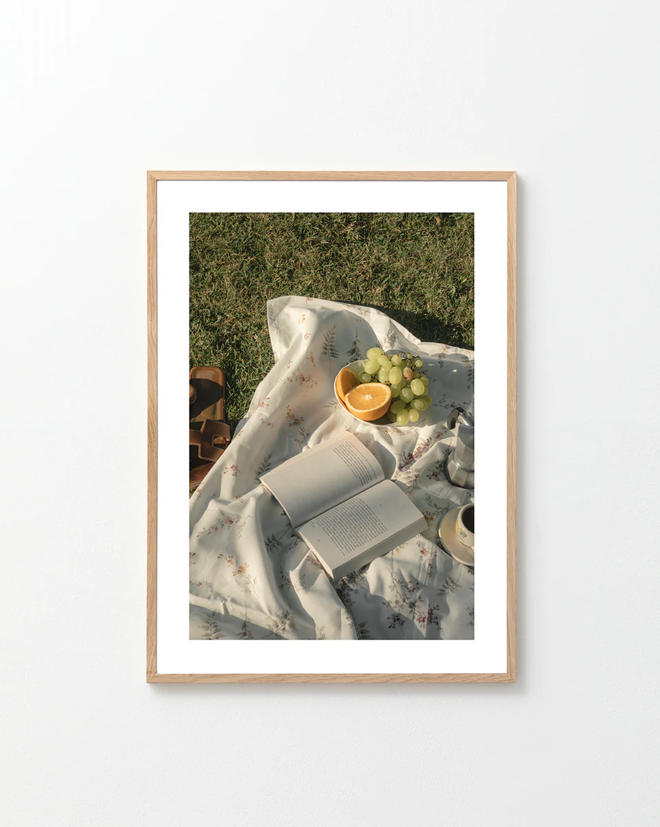 The Tuscan Summer Poster captures a serene picnic on lush grass with a floral blanket cradling an open book, sliced oranges, grapes, and a cup. Brown sandals add to the relaxed outdoor vibe, perfect for inviting moments under the sky.