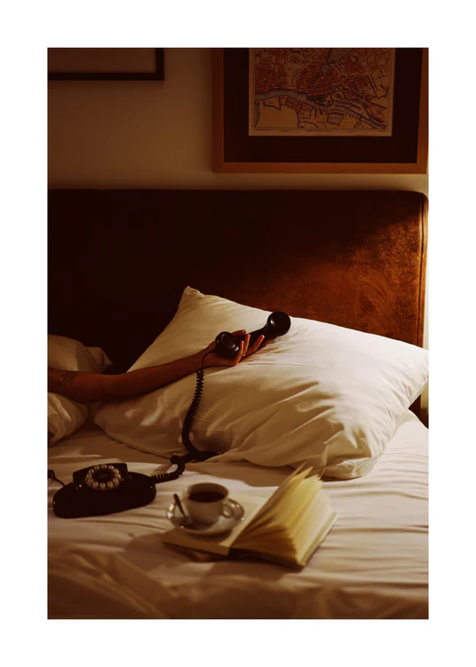 In a dimly lit bedroom, a person relaxes on a bed, vintage telephone in hand. Nearby, an open book and coffee sit invitingly. Above the headboard, the Do Not Disturb Poster adds elegance and nostalgia to the scene.