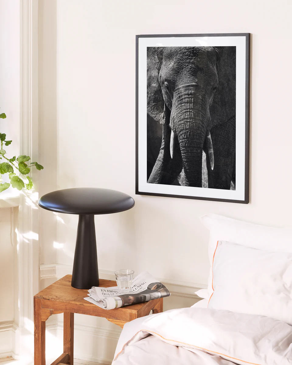 A cozy bedroom corner has a wooden stool holding a glass of water and a magazine near the bed. Nearby, theres a modern black side table, and the cream-colored wall displays the Gentle Authority poster featuring an elephant, with a plant peeking in from the window.