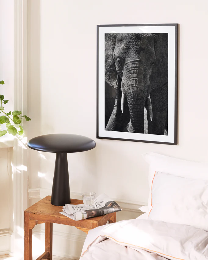 A cozy bedroom corner has a wooden stool holding a glass of water and a magazine near the bed. Nearby, theres a modern black side table, and the cream-colored wall displays the Gentle Authority poster featuring an elephant, with a plant peeking in from the window.