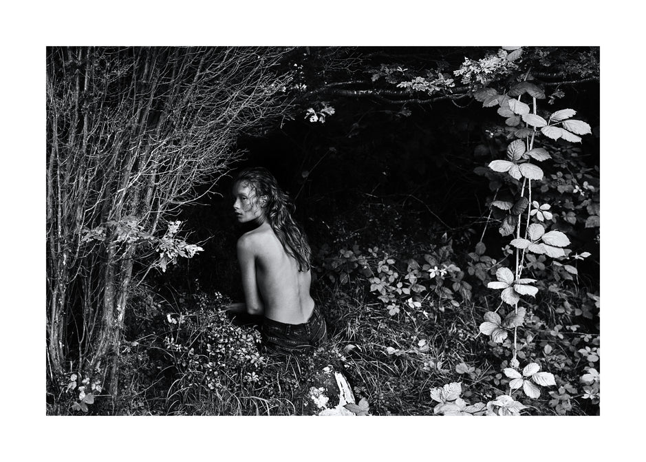 Woman sitting alone in a ditch surrounded by plants and bushes.