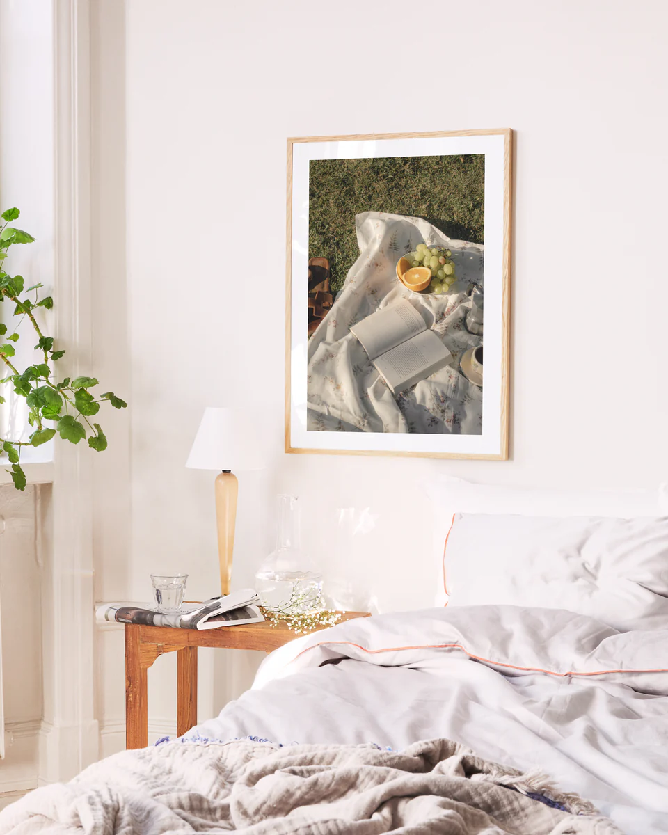 A cozy bedroom features a neatly made bed with white linens and a light brown blanket. On the wooden nightstand are books, a lamp, and a decorative plant. Above this serene setup, the Tuscan Summer Poster depicting a sunny picnic adds charm.