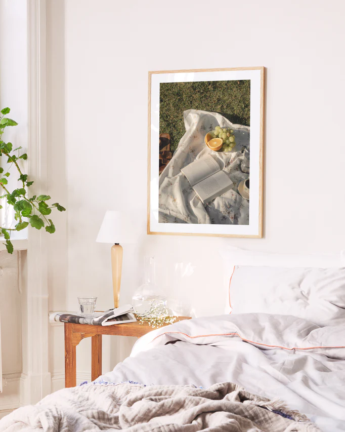 A cozy bedroom features a neatly made bed with white linens and a light brown blanket. On the wooden nightstand are books, a lamp, and a decorative plant. Above this serene setup, the Tuscan Summer Poster depicting a sunny picnic adds charm.