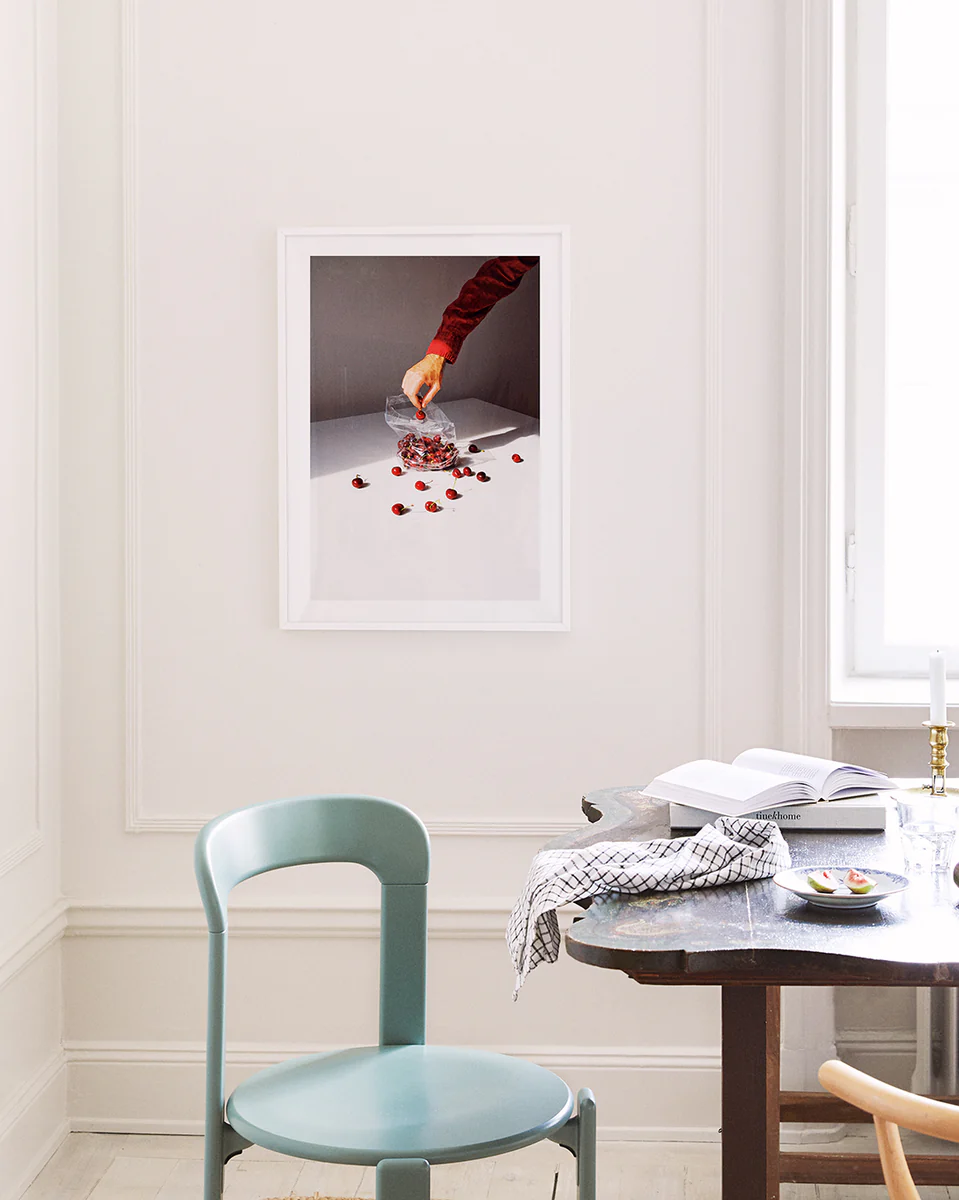 A minimalistic room features a teal chair and dark wooden table. The Summer Treats poster on the wall displays a hand with a red sleeve holding a bouquet above white, scattered with red petals. Natural light streams through a window on the right.