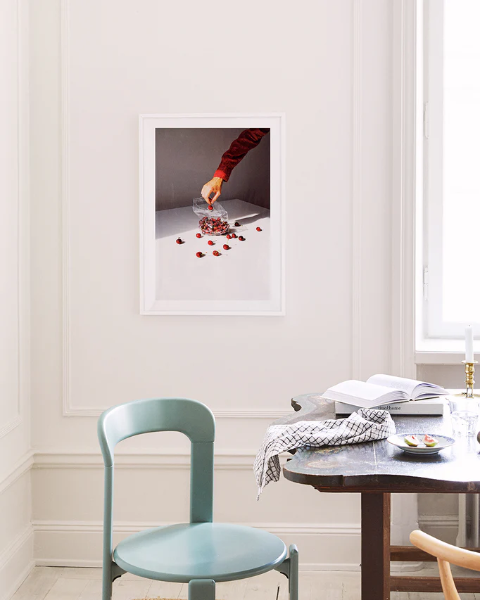 A minimalistic room features a teal chair and dark wooden table. The Summer Treats poster on the wall displays a hand with a red sleeve holding a bouquet above white, scattered with red petals. Natural light streams through a window on the right.