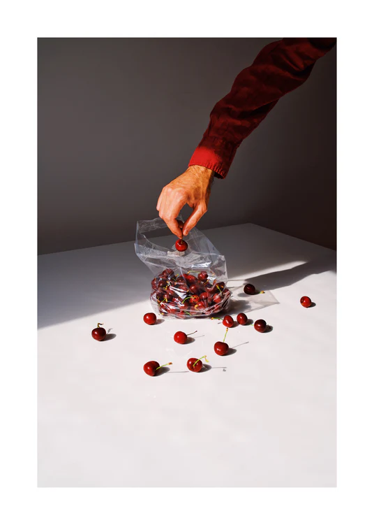A hand reaches into a clear bag filled with cherries, scattering several outside on a white surface. The sharp shadows highlight the rich red of the cherries in this Summer Treats Poster.