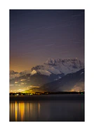 Star strewn sky above a city located next to a lake in the mountains.
