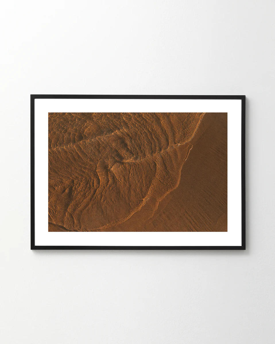 The Tide Poster, featuring brown sand patterns like moon-influenced tides, creates an abstract, earthy design with texture and shadows as it hangs on a white wall.