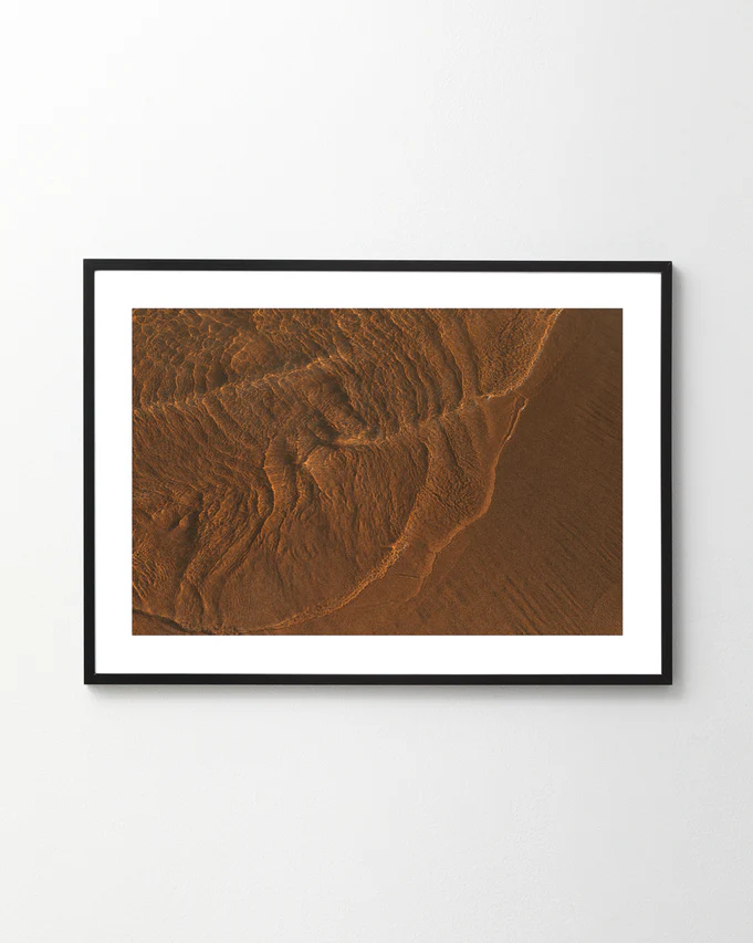 The Tide Poster, featuring brown sand patterns like moon-influenced tides, creates an abstract, earthy design with texture and shadows as it hangs on a white wall.
