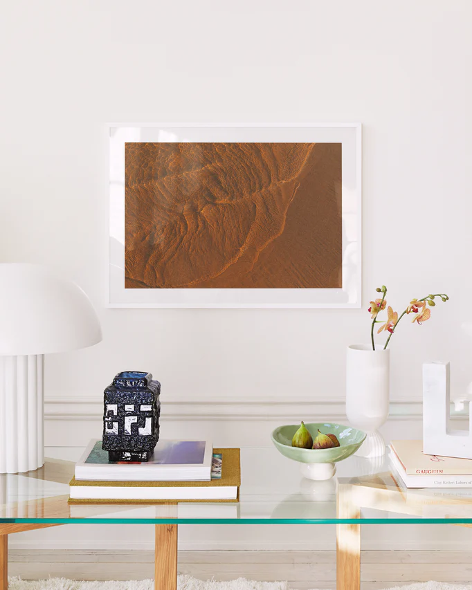 In a modern living room lit by moonlight, a glass coffee table holds books, a decorative blue vase, and green figs. A white lamp and an orchid adorn the table while the Tide Poster, depicting a desert landscape, hangs on the white wall.