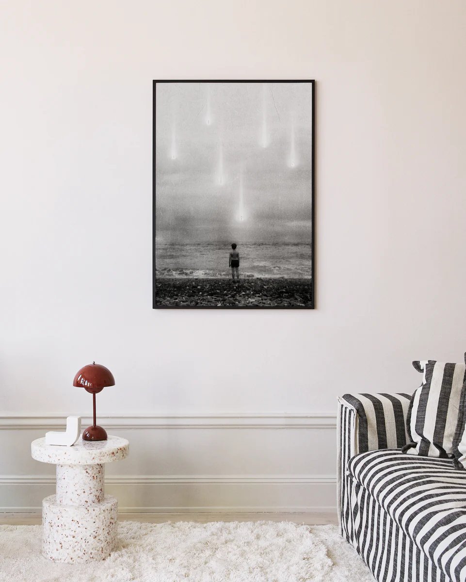 A modern living room showcases the Doubts Poster, a black-and-white photograph of a child facing the ocean with sky lights. The room features a striped sofa, small terrazzo table, and red lamp.