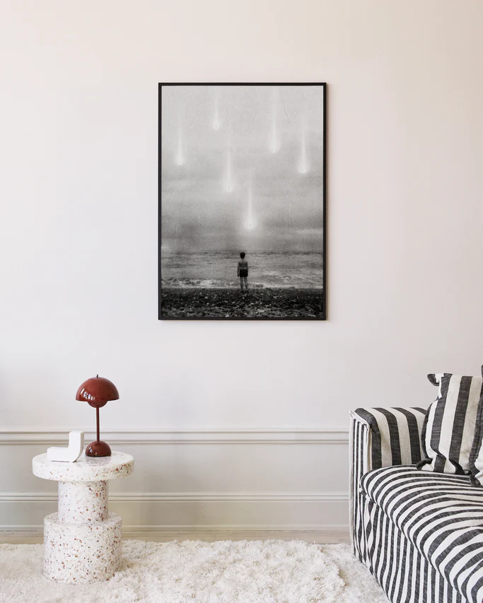 A modern living room showcases the Doubts Poster, a black-and-white photograph of a child facing the ocean with sky lights. The room features a striped sofa, small terrazzo table, and red lamp.