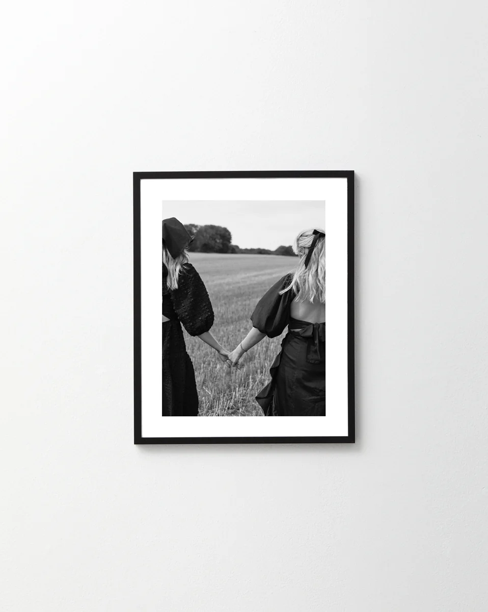 The Stand By Me, But Make It Fashion Poster features a black and white image of two friends with long hair, wearing dresses and hats, holding hands as they embark on an adventure in a sunny open field.