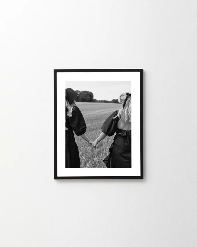 The Stand By Me, But Make It Fashion Poster features a black and white image of two friends with long hair, wearing dresses and hats, holding hands as they embark on an adventure in a sunny open field.