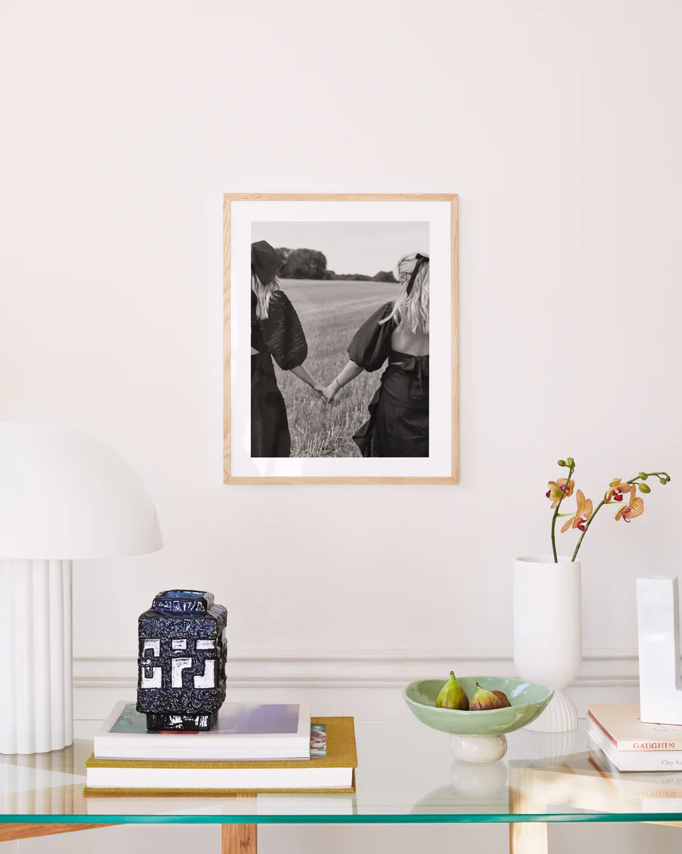 In a well-lit room, a glass table displays books, a patterned candle, a bowl with figs, and an orchid vase. A Stand By Me, But Make It Fashion poster of two friends in an open field hangs on the wall. A modern white lamp stands close by.