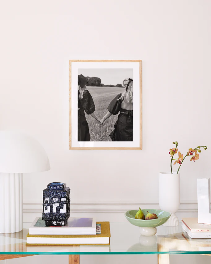 In a well-lit room, a glass table displays books, a patterned candle, a bowl with figs, and an orchid vase. A Stand By Me, But Make It Fashion poster of two friends in an open field hangs on the wall. A modern white lamp stands close by.