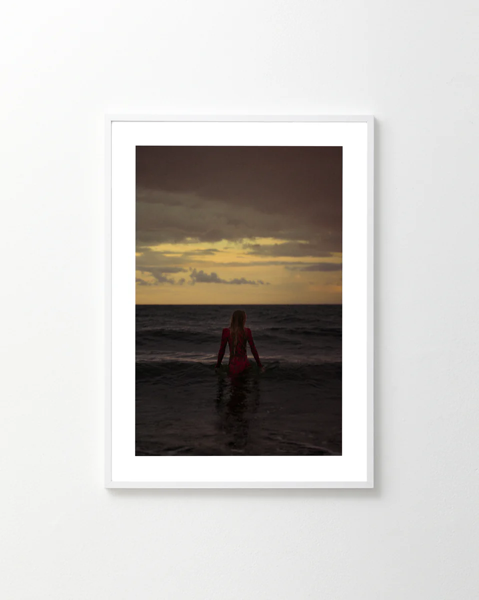 The Seizing the Last Moments of the Day Poster showcases a person standing in the ocean at sunset, with a dramatic, cloud-filled sky. Facing the horizon and partly submerged, theyre illuminated by warm light on the sea, evoking FOMO.