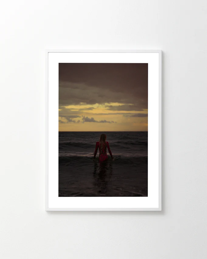 The Seizing the Last Moments of the Day Poster showcases a person standing in the ocean at sunset, with a dramatic, cloud-filled sky. Facing the horizon and partly submerged, theyre illuminated by warm light on the sea, evoking FOMO.