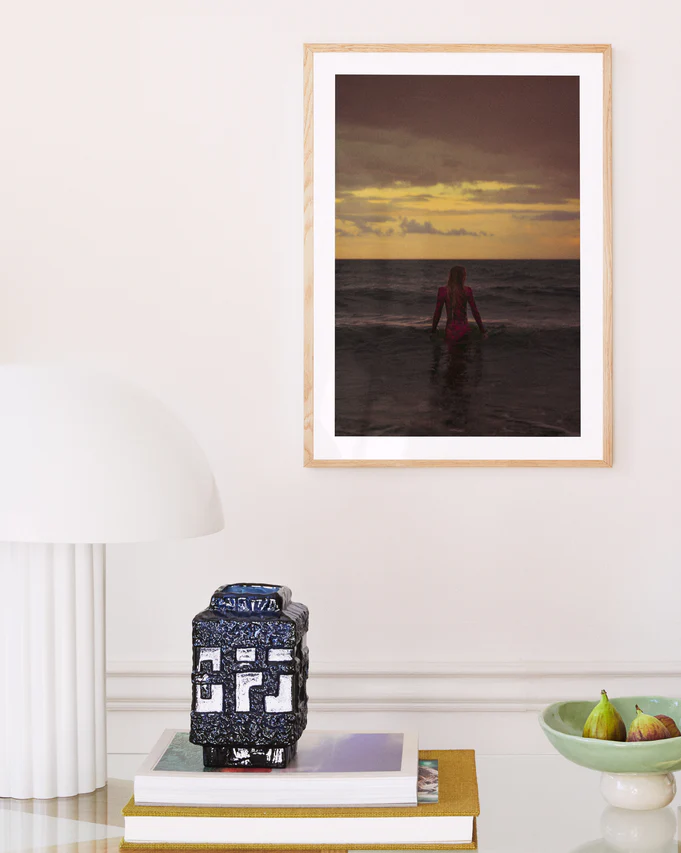 The Seizing the Last Moments of the Day poster adorns a light-toned wall above a peaceful arrangement. Below on the shelf, a white lamp, blue decorative vase, and fig-filled bowl contribute to an inviting atmosphere that reduces FOMO and helps you relax before bed.