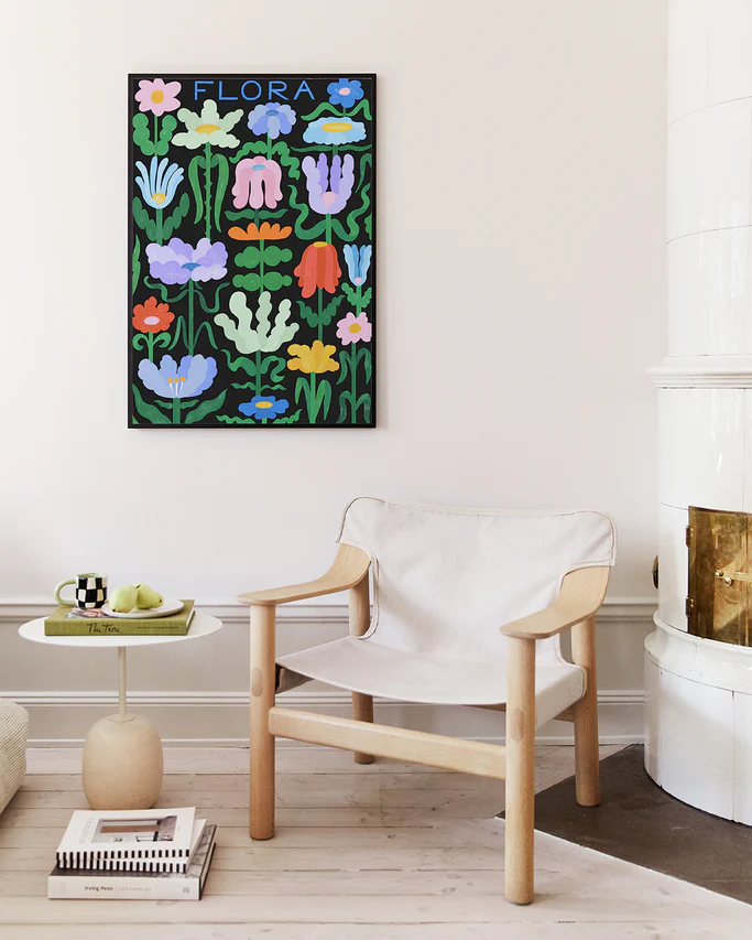 A cozy room features a beige chair and a small table with a checkered cup, plate, and green apples. The wall above a white fireplace is adorned with Flora on Black poster with colorful floral artwork and handwritten typography. Books rest on the light wood floor nearby.