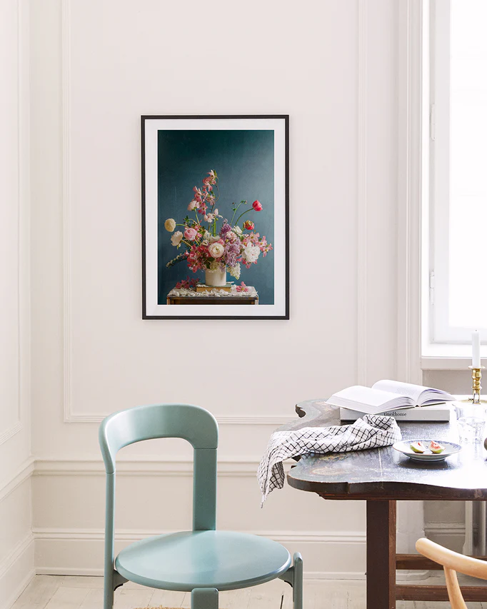 A minimalist room features a teal chair, dark wood table, and an open book. Near the window with soft light is the Spring Ballet Poster, showcasing a vase of flowers as colorful wall art hanging on a white wall.