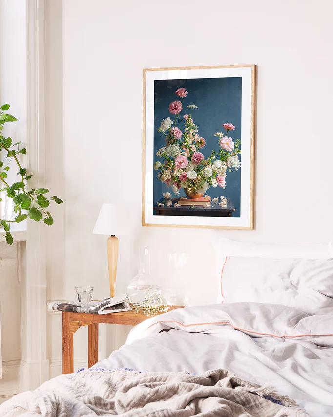 A cozy bedroom features a neatly made bed with white bedding and a throw. A wooden side table holds a lamp, books, and a carafe. Above the bed, the Midsummer poster adorns the wall while a leafy plant sits by the window.