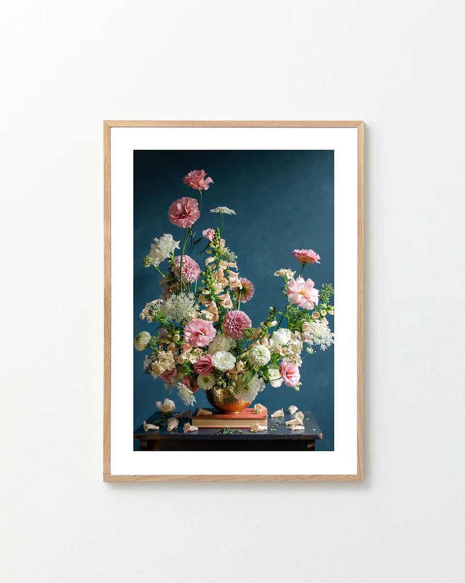 The Midsummer Poster showcases a framed photograph of a pink and white floral arrangement with greenery on a dark wooden table, set against a dark blue background. Some petals are scattered around, and the frame features a light wood finish.