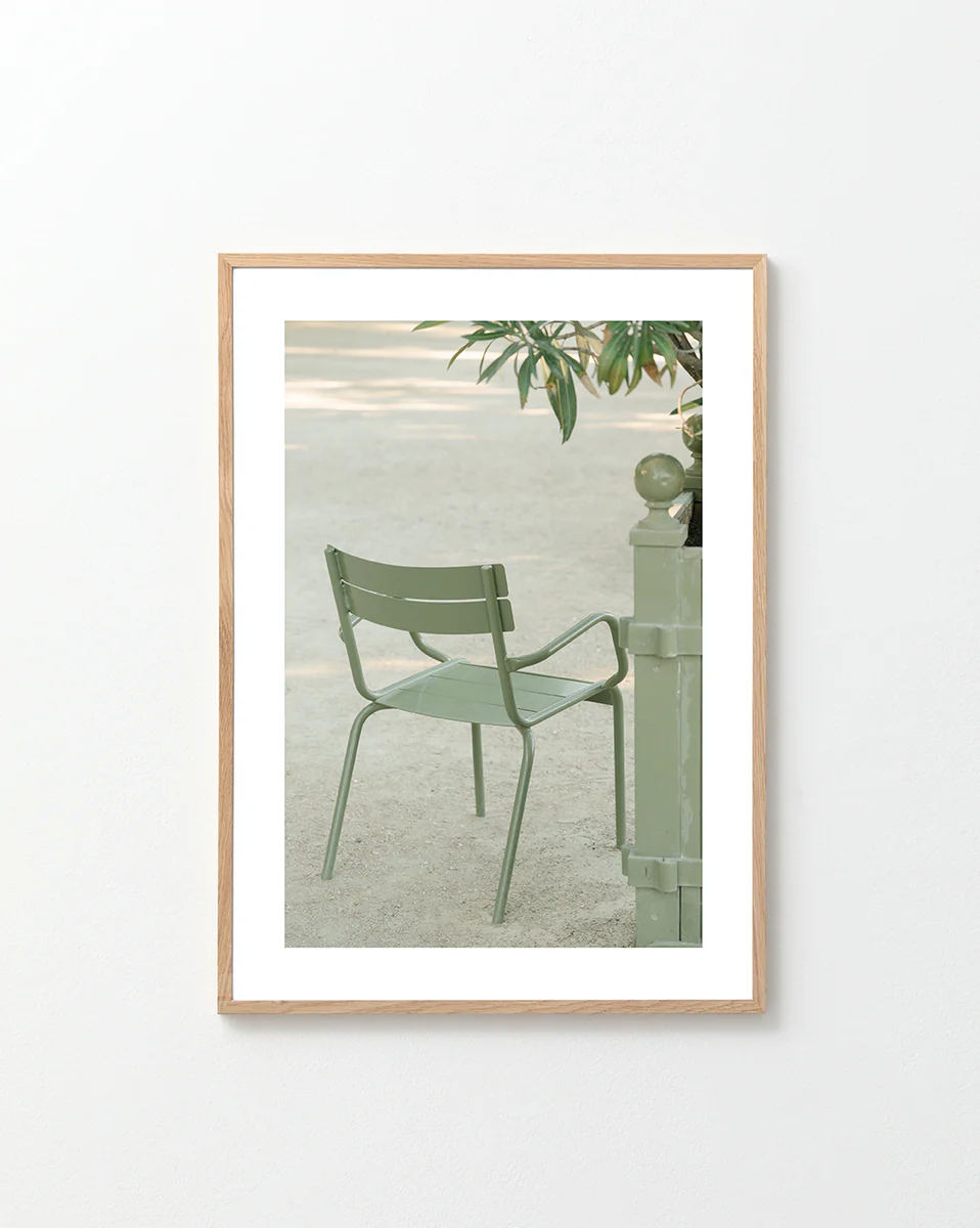 The Asseyez-vous Poster features a light green metal chair beside a matching post with overhanging branches against a sandy outdoor background. Its framed in light wood and displayed on a white wall.