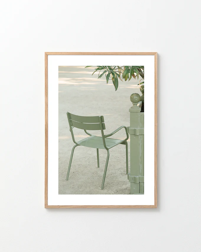 The Asseyez-vous Poster features a light green metal chair beside a matching post with overhanging branches against a sandy outdoor background. Its framed in light wood and displayed on a white wall.