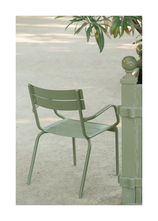 On a sandy surface under soft sunlight, an Asseyez-vous Poster is set beside a decorative post and leafy plant, creating a serene and inviting outdoor scene.