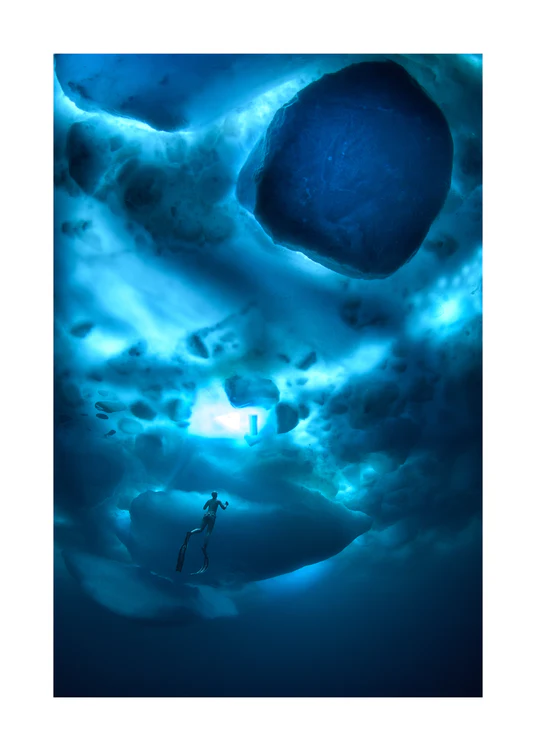 A diver swimming in the deep blue ocean underneath a surface made from ice.