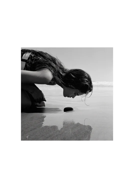 Woman sitting in the shallows of a beach looking down on a small black rock.