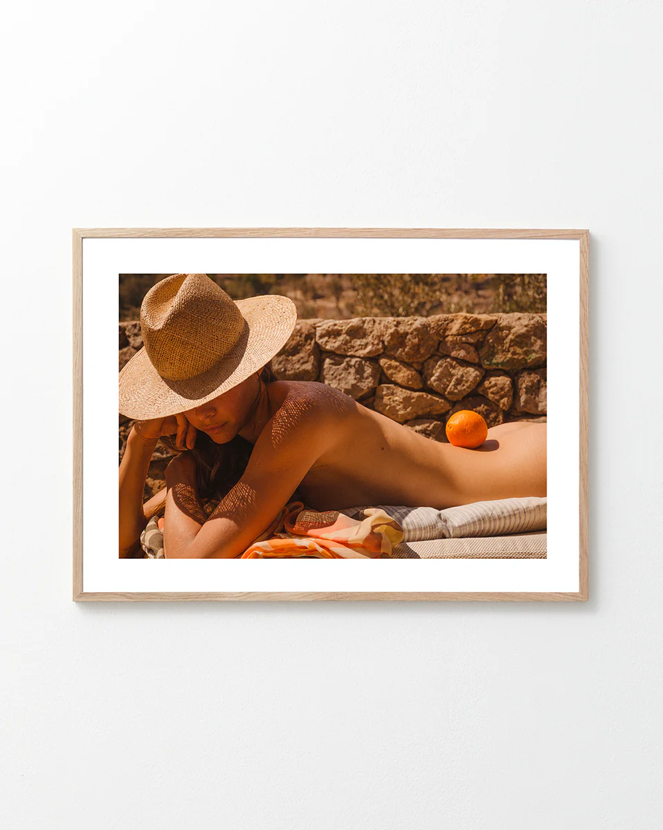 Naked woman on a sun bed wearing a hat and balancing an orange on her back.