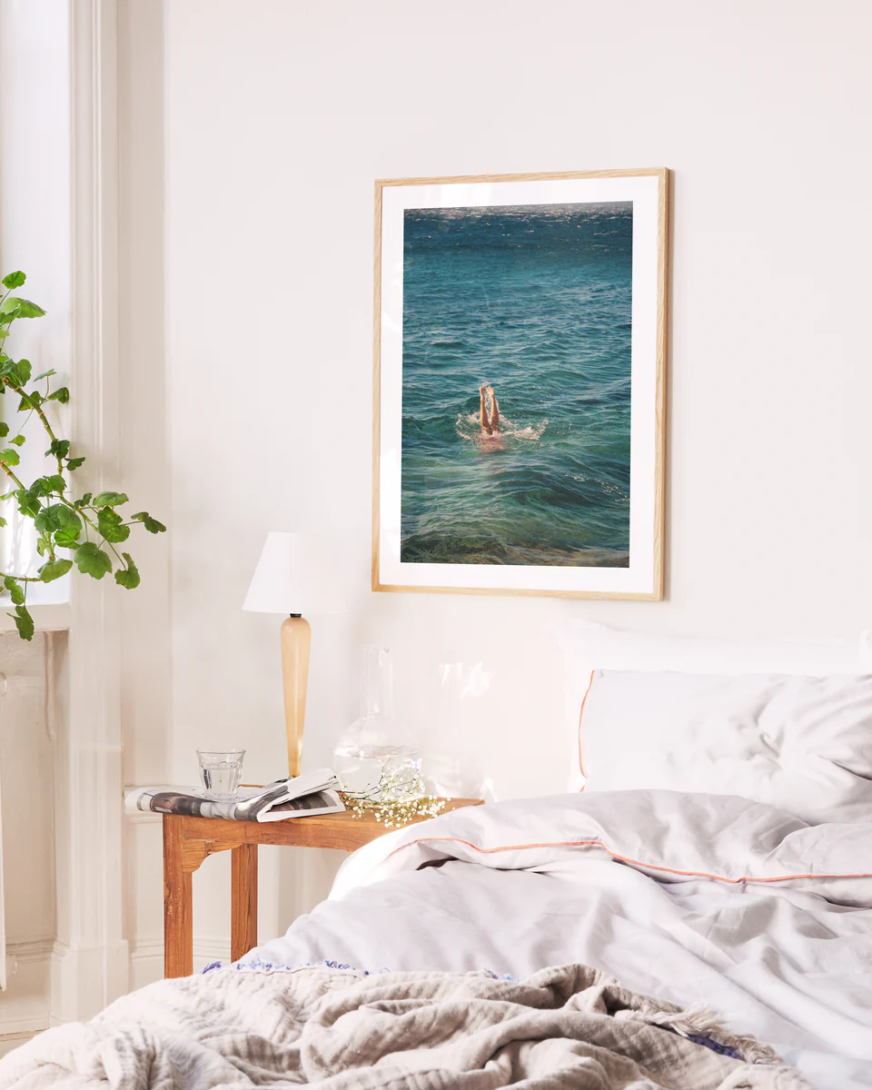 A cozy bedroom features a white bed and wooden nightstand. Above the bed hangs the Morning Dip Poster by Lavinia Cernau, depicting a swimmer in the ocean. By the tall window, a potted plant adds life, while a lamp, books, and glass vase adorn the nightstand.
