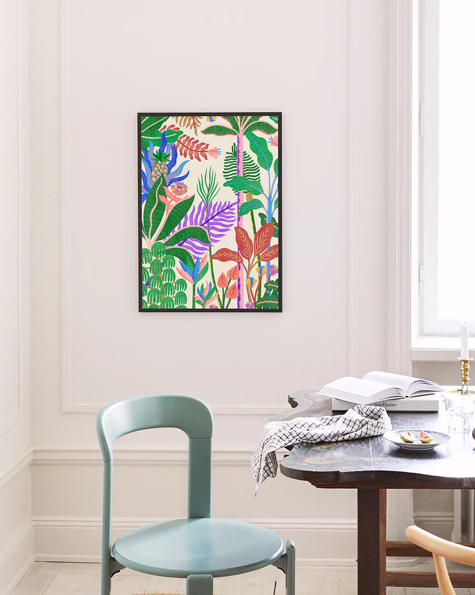 The Jungle by Day Poster by Manne Jalilian showcases lively botanical artwork on a white wall. Below, a wooden table with an open notebook and checkered cloth complements the setting. A light blue chair is nearby, illuminated by sunlight streaming through the window.
