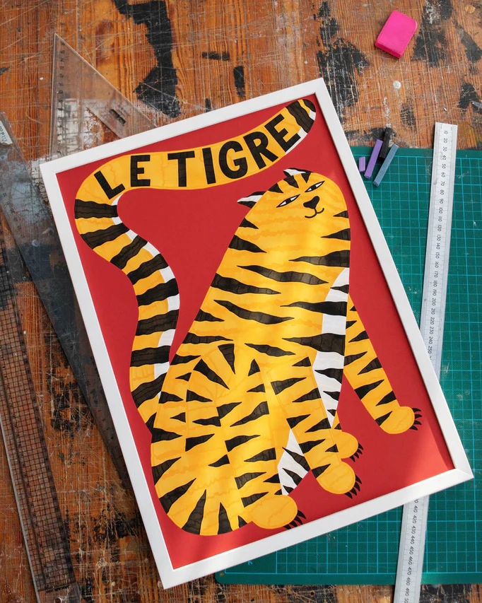 The Le Tigre Red Poster by Manne Jalilian shows a stylized orange tiger with yellow and black stripes on a vivid red background. Its displayed on a wooden table surrounded by rulers, a cutting mat, and chalk pieces.