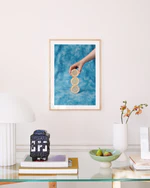 A minimalist room displays a framed Sour Poster, featuring a hand holding a lemon slice against a blue background. Below, on the glass table, are a modern lamp, decorative jar, books, a bowl with figs, and an orchid in a vase.