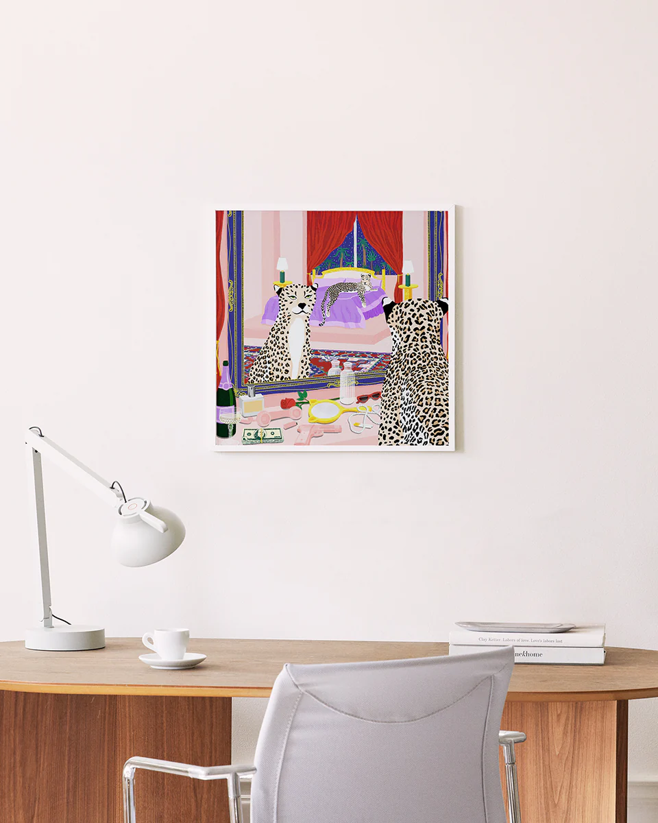 A minimalist workspace with a wooden desk includes a white lamp, books, and a cup; the chair is pushed in. Above it hangs the colorful Bonafyde Poster showing two leopards gazing into a mirror in a lavish room.