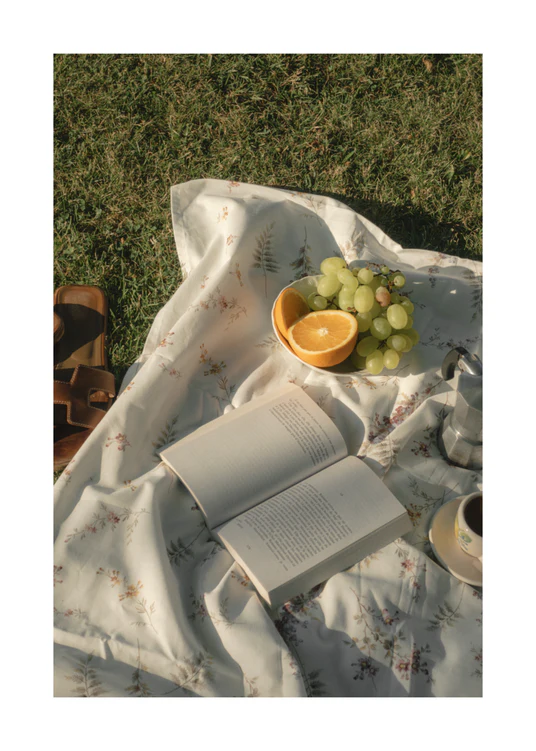 The Tuscan Summer Poster showcases a picnic scene on a floral blanket with an open book, juicy green grapes, a vibrant orange, an elegant leather bag, and a ceramic cup on lush grass. Soft lighting creates an inviting atmosphere under the suns warm glow.