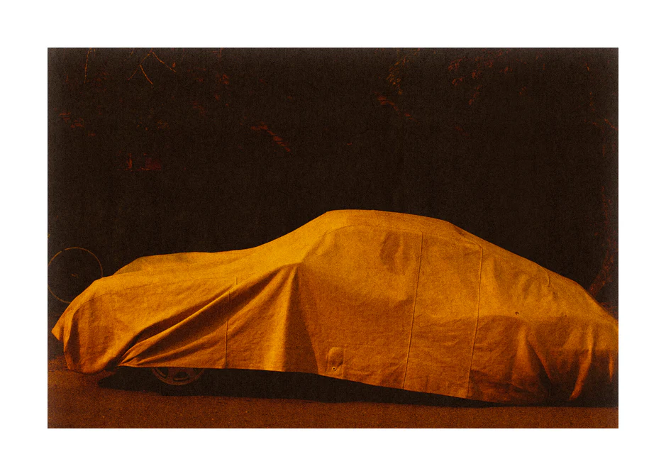 Car covered in a white sheet turned orange underneath the light of a streetlamp.