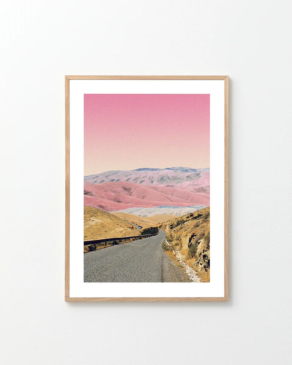 The Rosé Road Poster features a framed photograph of a winding road through hills in earthy tones under a surreal pink sky. The light wood frame stands out against a plain white wall.