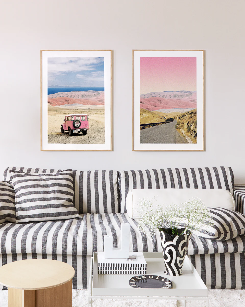 A stylish living room with a black-and-white striped sofa and matching cushion conjures fairy-tale charm. Two Rosé Road Posters on the wall act as windows to another world, while a white coffee table with a black-and-white vase of flowers completes the enchanting scene.