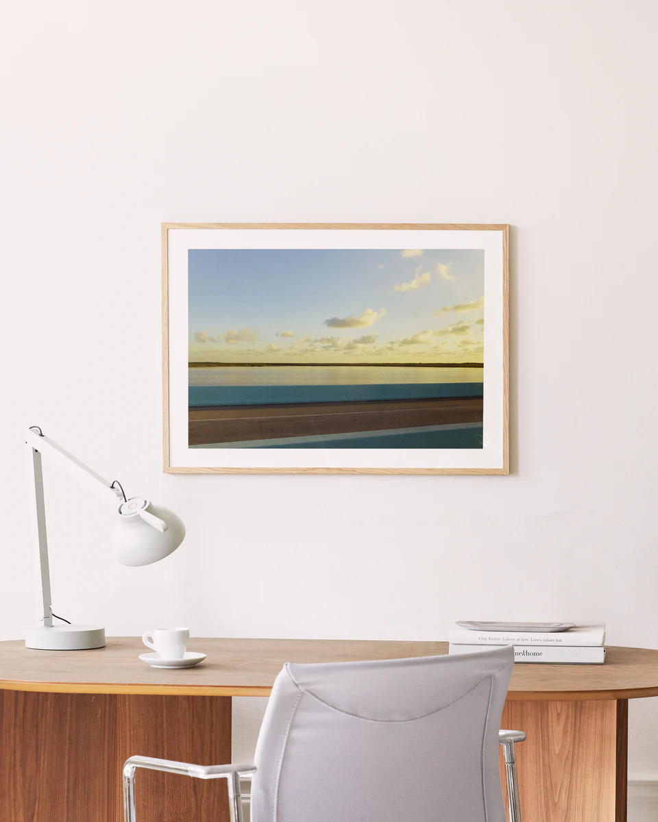 A minimalist wooden desk with a white lamp, cup, and books sits beside the road to creativity. In front is a light gray chair. Gracefully hanging above on the cream-colored wall is a wooden-framed Water Split Poster depicting clouds and water.