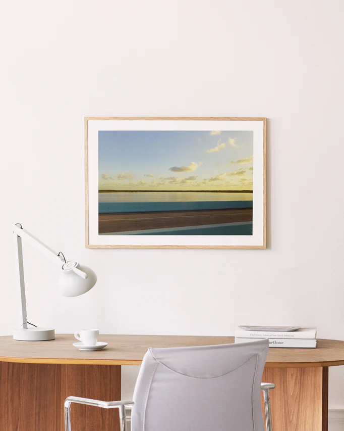 A minimalist wooden desk with a white lamp, cup, and books sits beside the road to creativity. In front is a light gray chair. Gracefully hanging above on the cream-colored wall is a wooden-framed Water Split Poster depicting clouds and water.