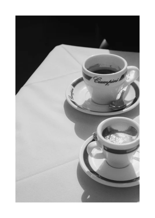 A nostalgic black and white photo features two coffee cups on saucers with spoons, set against a white tablecloth. This scene, resembling a vintage Rome café, mirrors the peaceful allure of classic Ciampini Poster designs with logo-adorned ceramics.