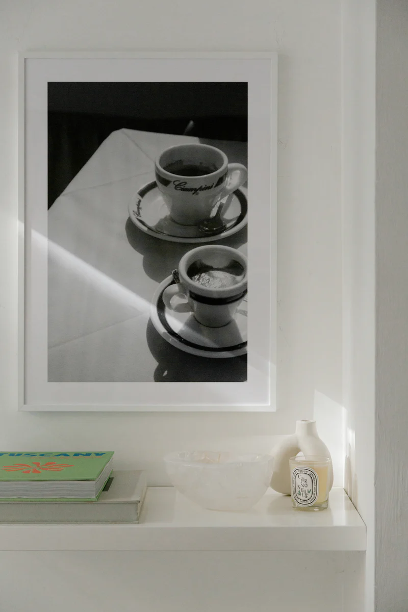 The Ciampini Poster, a framed black and white photo reminiscent of vintage Rome, hangs above a white shelf. On the shelf are two books, a clear bowl, and a small candle jar. Sunlight softly illuminates the scene.