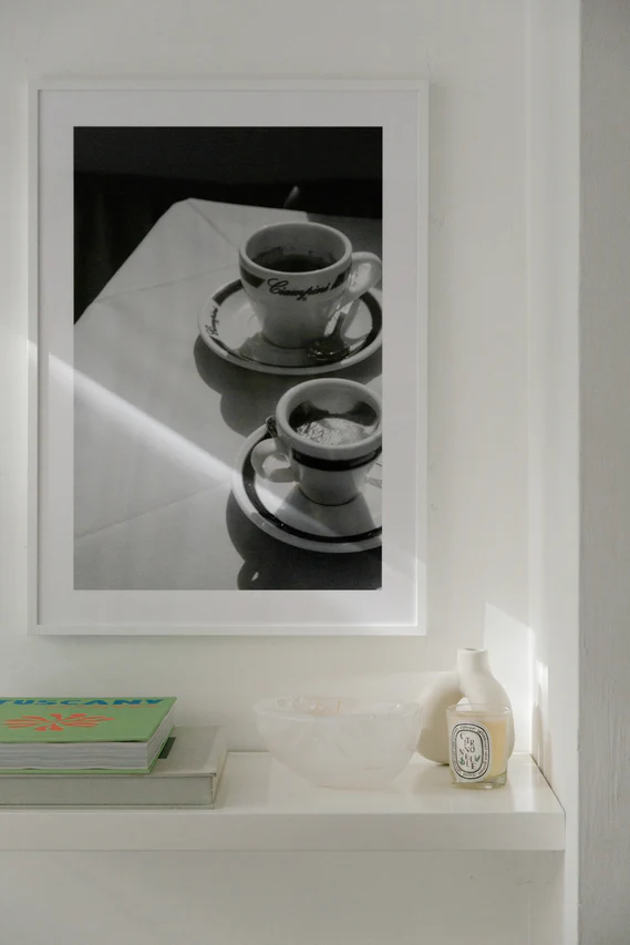 The Ciampini Poster, a framed black and white photo reminiscent of vintage Rome, hangs above a white shelf. On the shelf are two books, a clear bowl, and a small candle jar. Sunlight softly illuminates the scene.