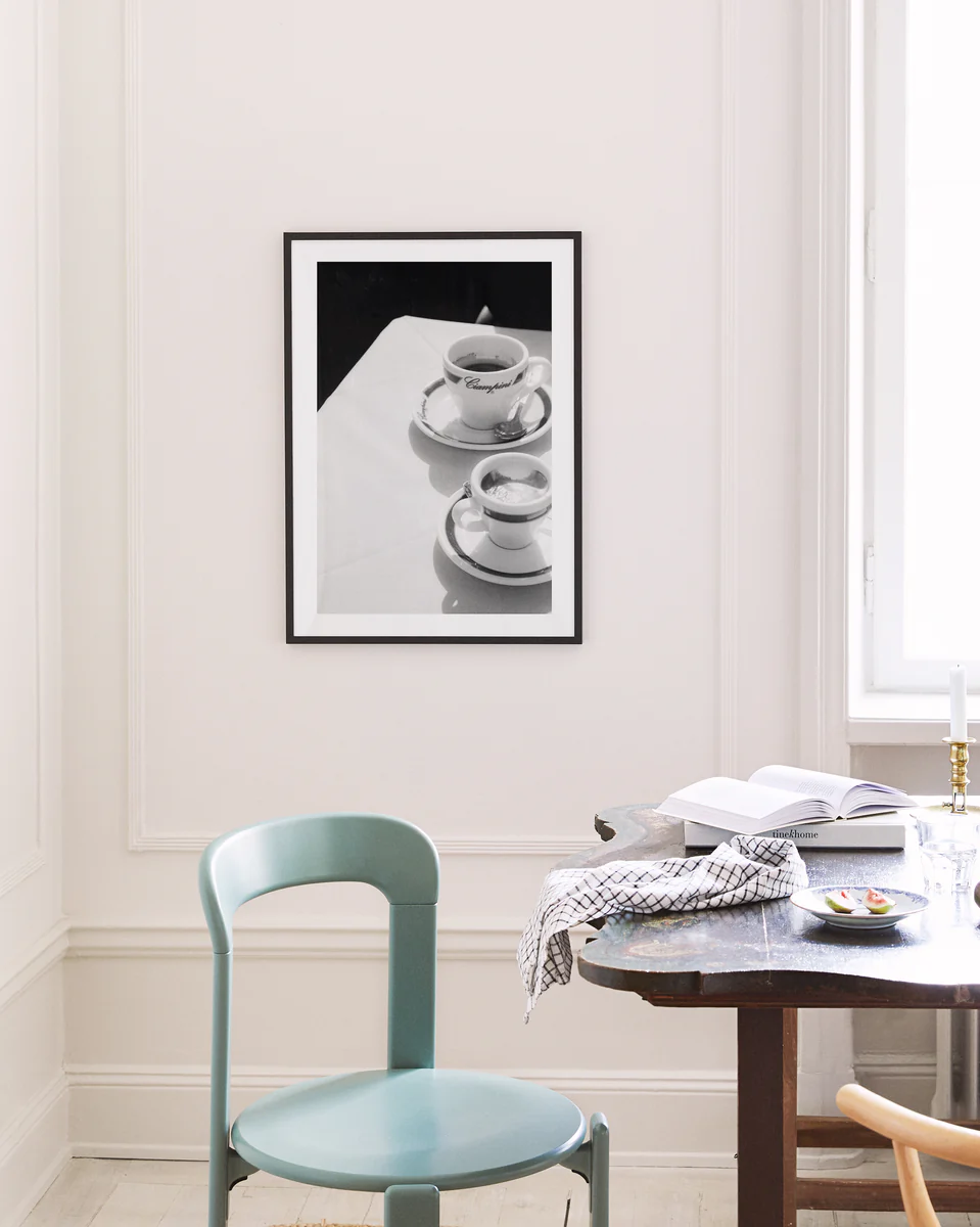 In a minimalist room adorned with white walls and subtle decor, the Ciampini Poster, showcasing a framed black-and-white photo of two coffee cups evoking vintage Rome, hangs above. A wooden table with an open book, plate, and checked cloth complements the light blue chair nearby.