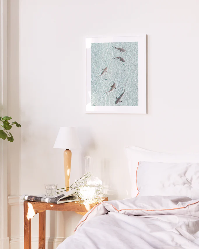 A bright bedroom showcases a neatly made bed with a white pillow and comforter. A small wooden table beside it bears a lamp, water glass, and book. Above the bed, the On Patrol poster adds calmness, while subtle background music suggests an adventurous undertone.