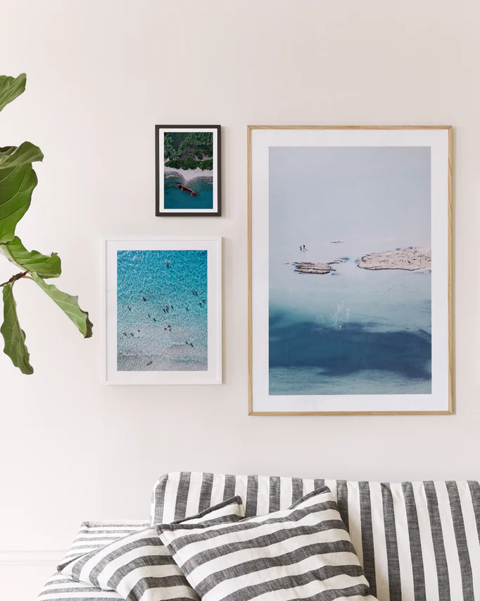 A beige wall showcases three framed photos: the Eavesdropping IX Poster depicting a beach scene with swimmers, another showing a rocky island in turquoise water, and a smaller serene beach view. A striped couch and leafy plant create an inviting space perfect for relaxation after a long day.